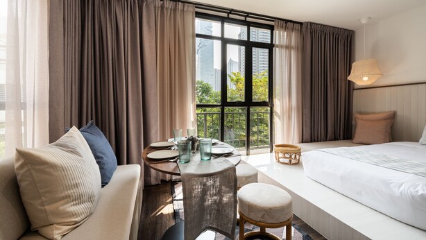 A hotel room with a large bed, a sofa, a small table with a vase, and a view of the city through large windows.