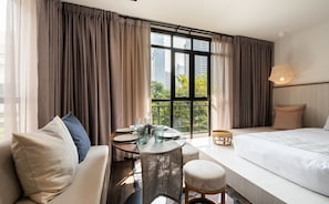 A hotel room with a large bed, a sofa, a small table with a vase, and a view of the city through large windows. - Warm Window Silom - Hostel (Bangkok)