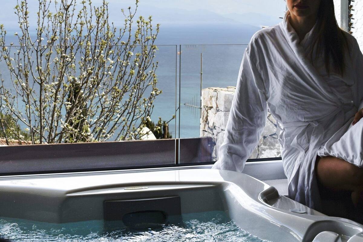 Iemand in een witte badjas zittend in een jacuzzi met uitzicht op de oceaan en een boom op de achtergrond.