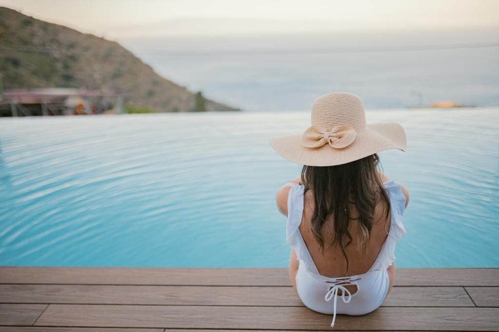 Eine Person sitzt auf einer Holzterrasse an einem Pool mit Blick auf das Meer und eine Hügelkette im Hintergrund.