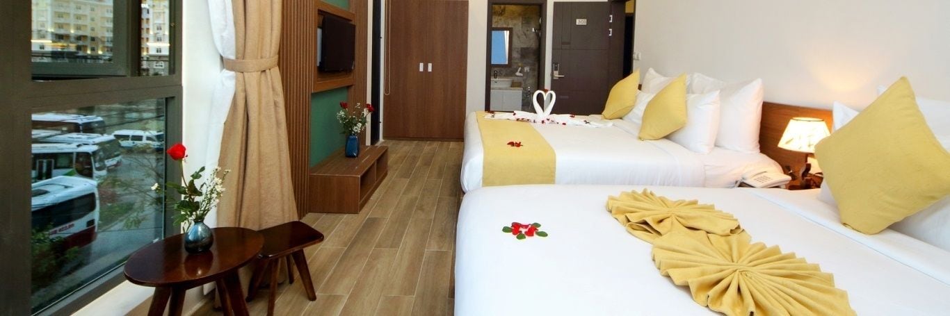 Habitación de hotel con dos camas, una mesita con un florero y vista a la ciudad a través de un ventanal grande.