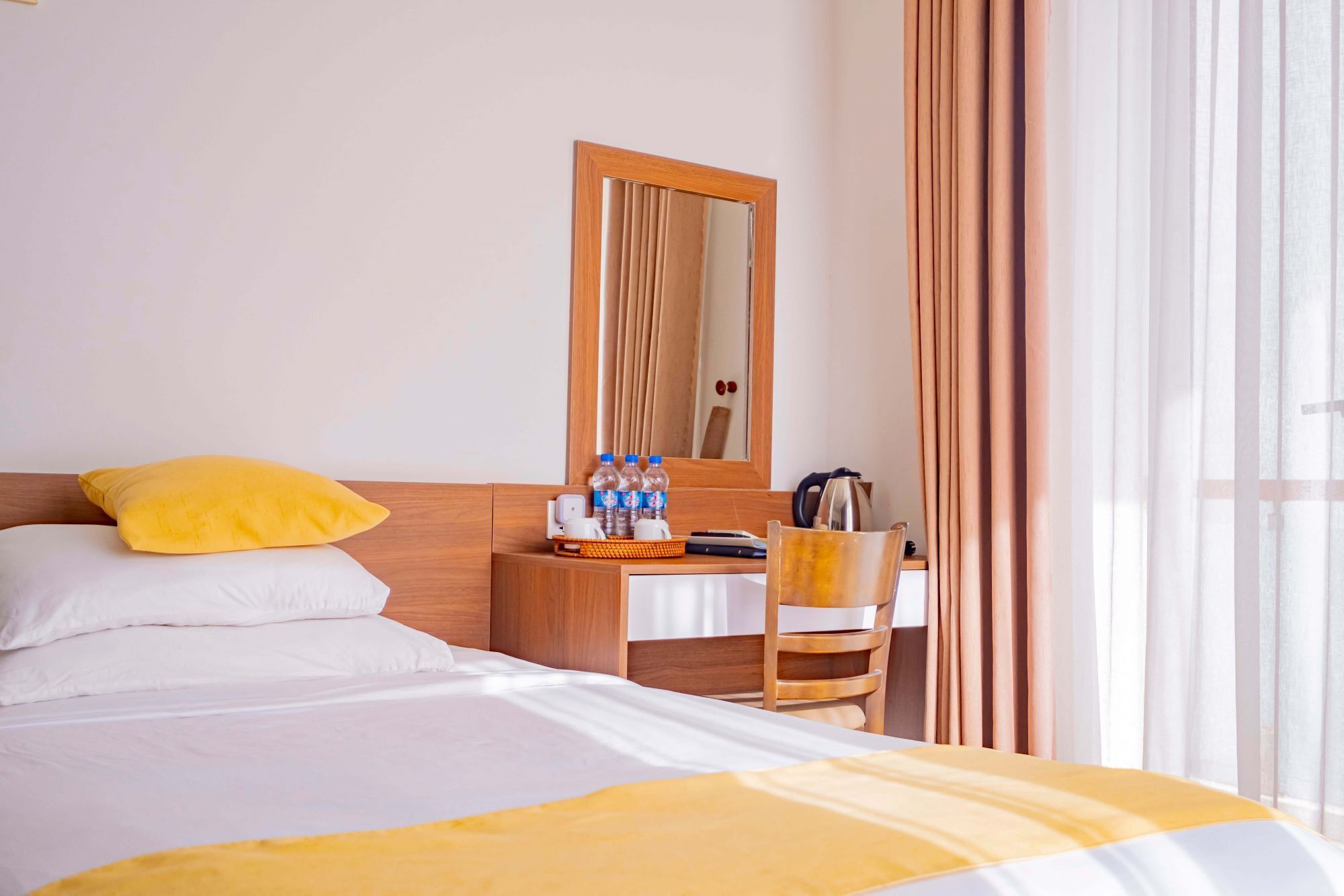A hotel room with a bed, a wooden nightstand with a mirror, a chair, and a window with curtains.