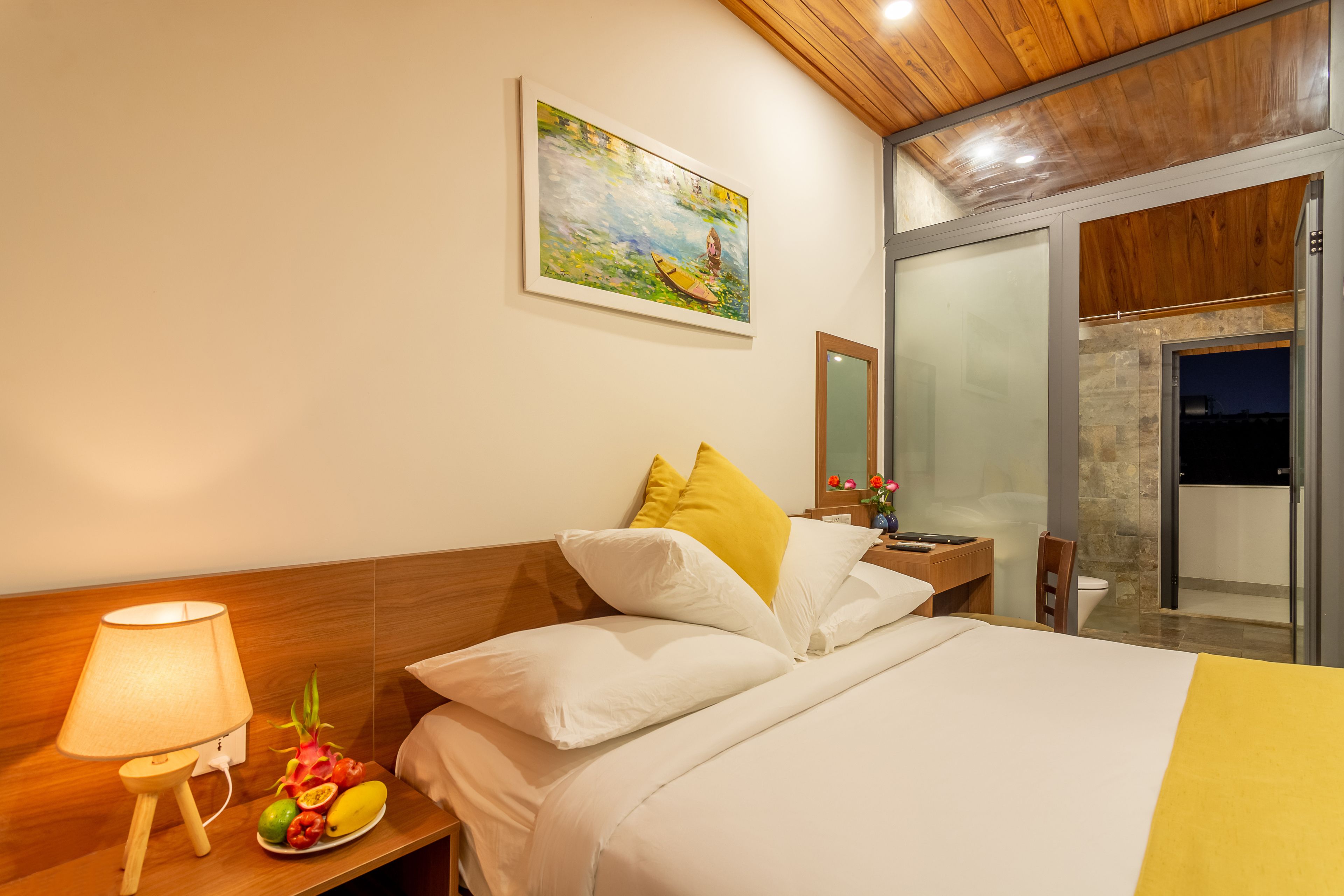 A hotel room with a bed, a nightstand with a lamp, a fruit plate, and a painting on the wall.