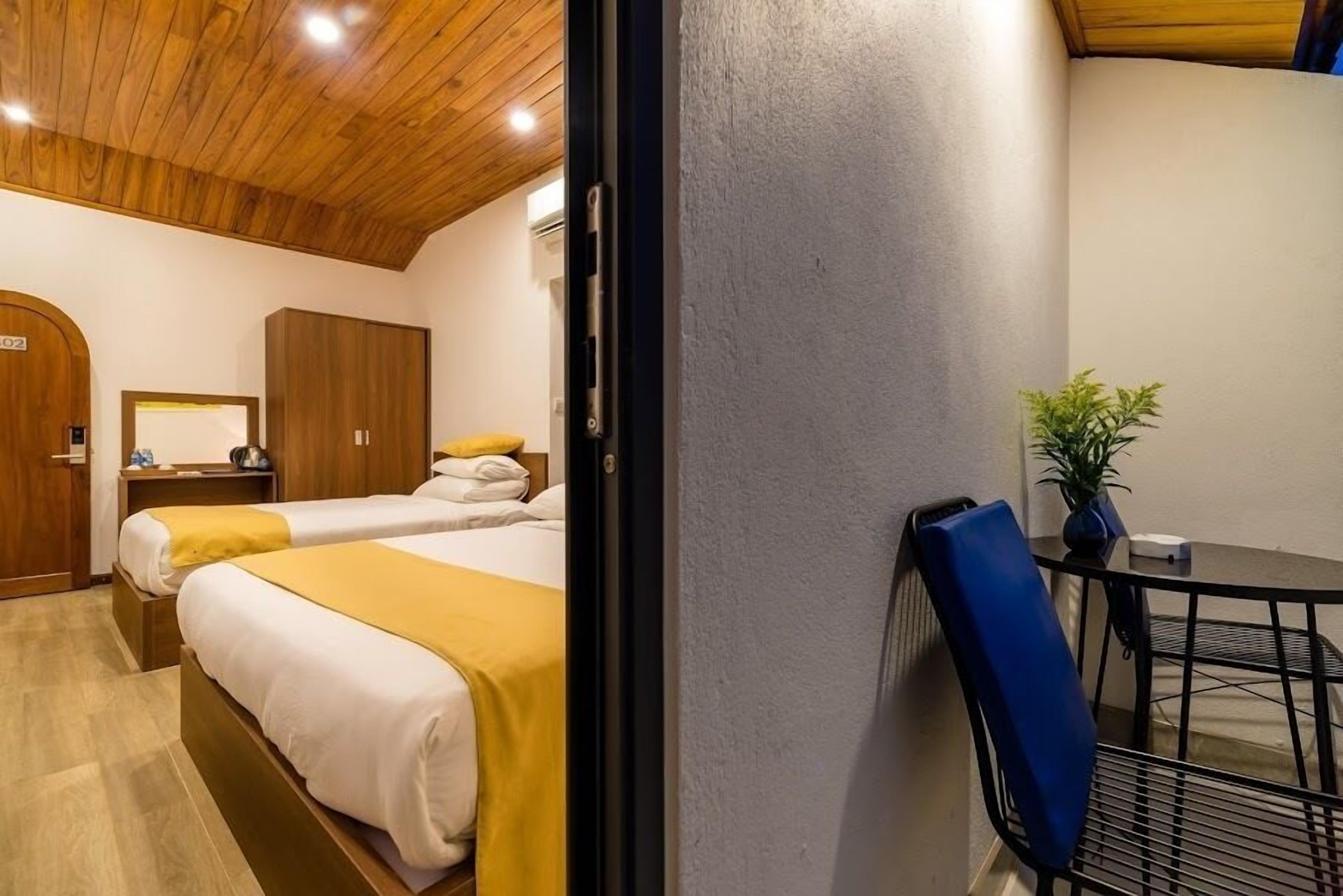 A hotel room with two beds, a wooden ceiling, a small table with a chair, and a potted plant.