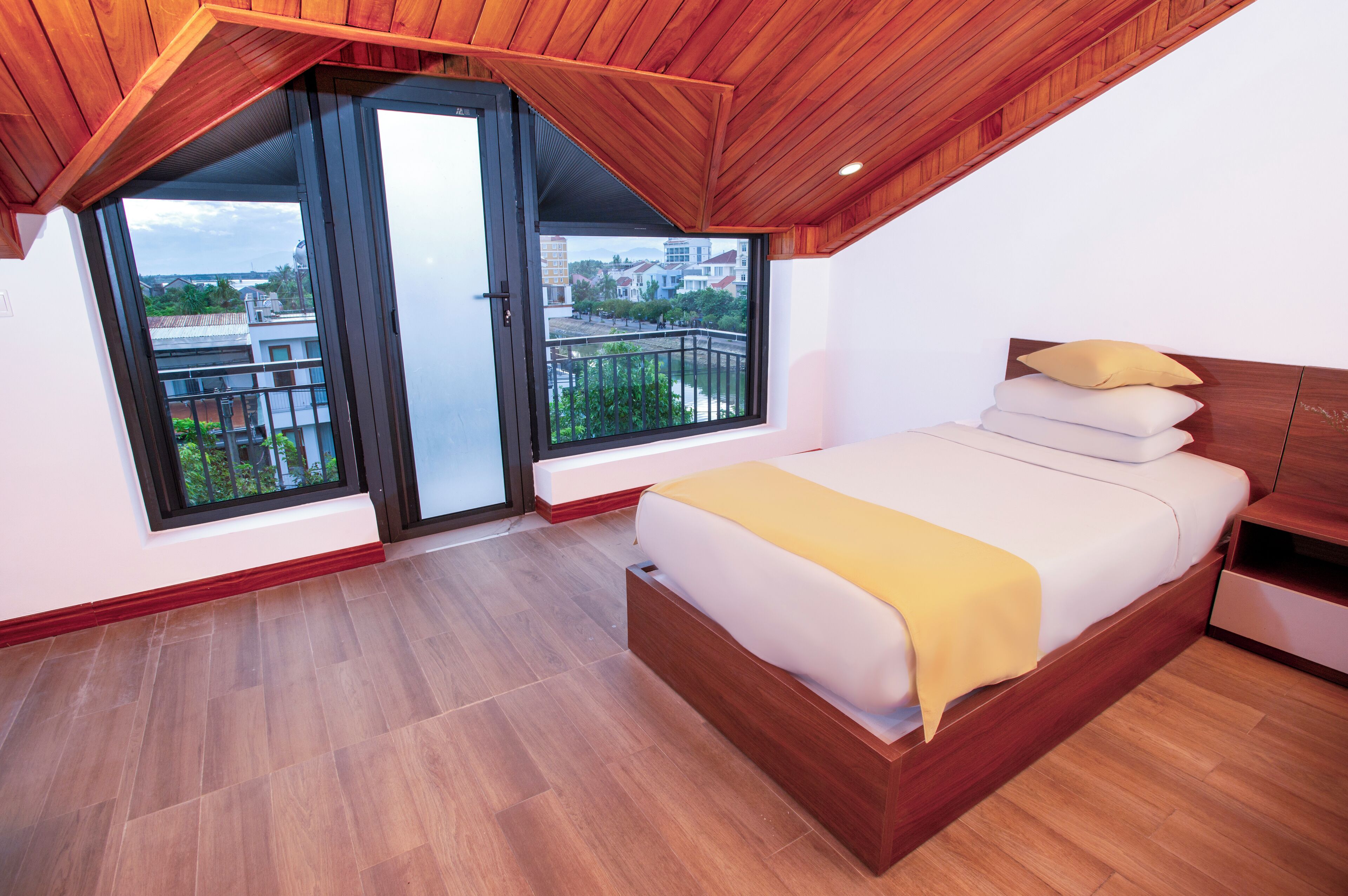 A bedroom with a wooden ceiling, a large bed with white bedding and a yellow throw, and a balcony with a view of buildings.