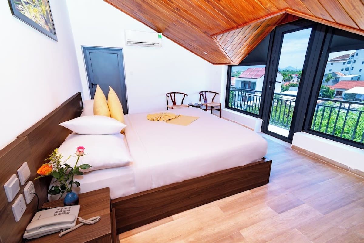 Ein modernes Hotelzimmer mit einem großen Bett, einem Holzkopfbrett, einem Nachttisch mit Telefon und Blick auf die Stadt durch große Fenster.