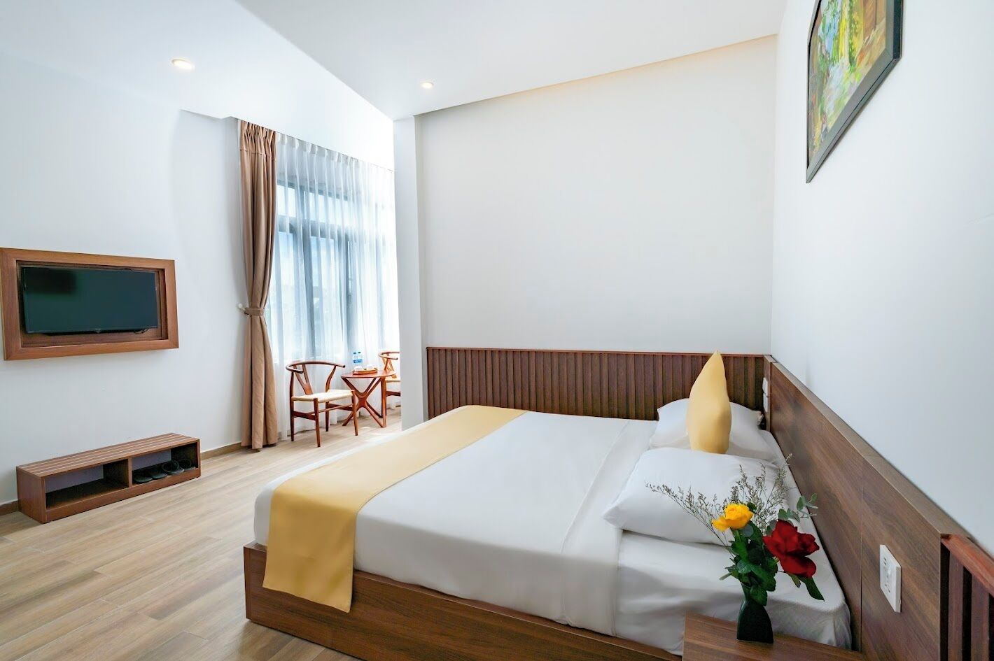 A hotel room with a bed, a TV mounted on the wall, a small table with chairs, and a window with curtains.