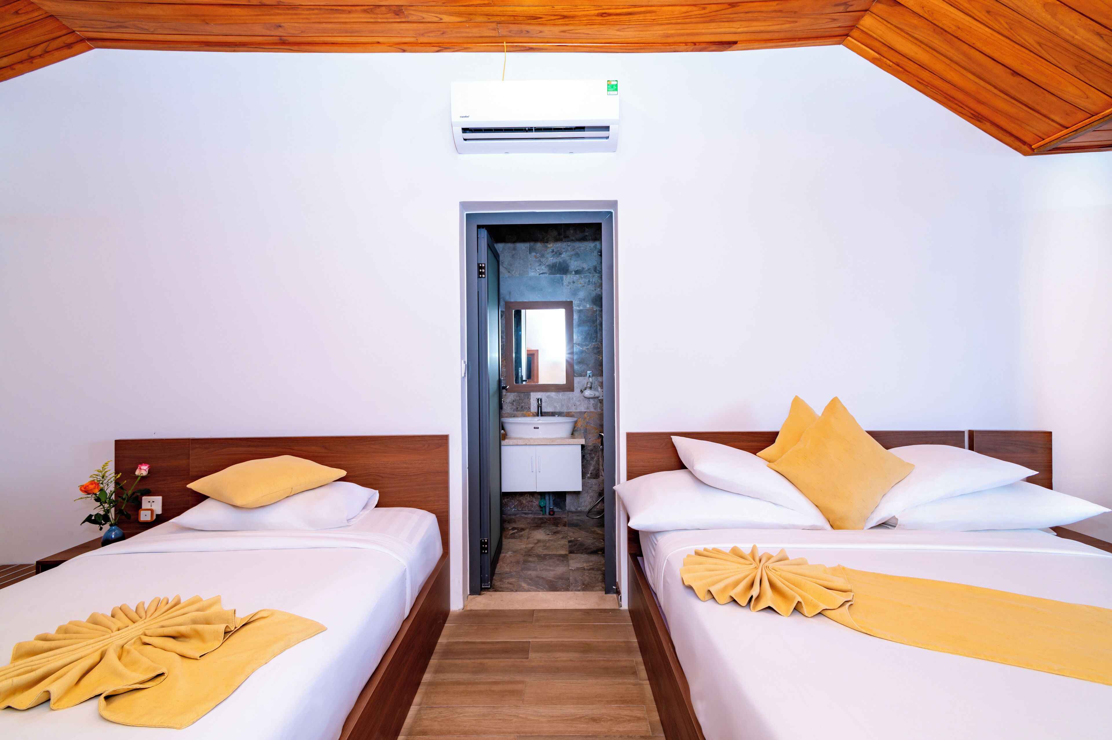 Une chambre d’hôtel avec deux lits, un plafond en bois, un appareil de climatisation et une salle de bain visible à travers une porte ouverte.