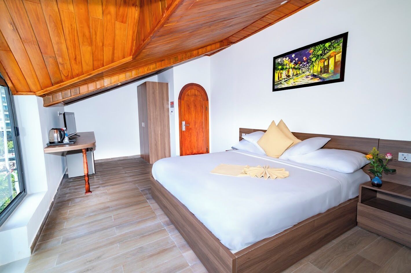 A modern bedroom with a wooden ceiling, a bed with white linens, a desk with a kettle, and a painting on the wall.