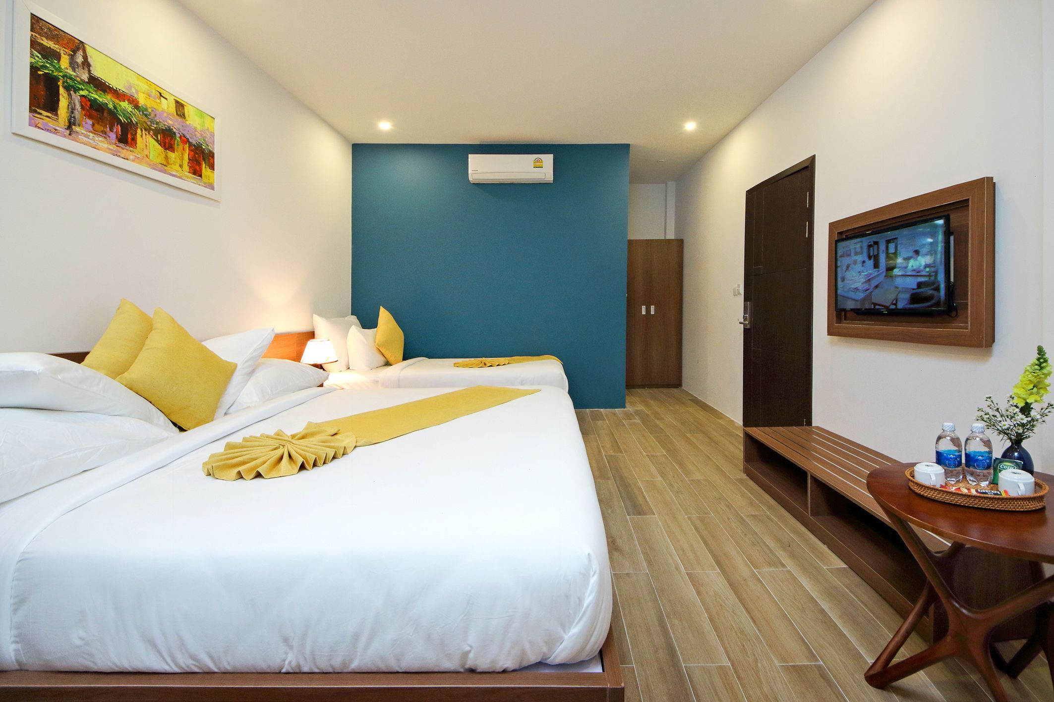 Een hotelkamer met een groot bed, een kleintje tafeltje met een vaas en flessen, een televisie aan de muur en een schilderij boven het bed.