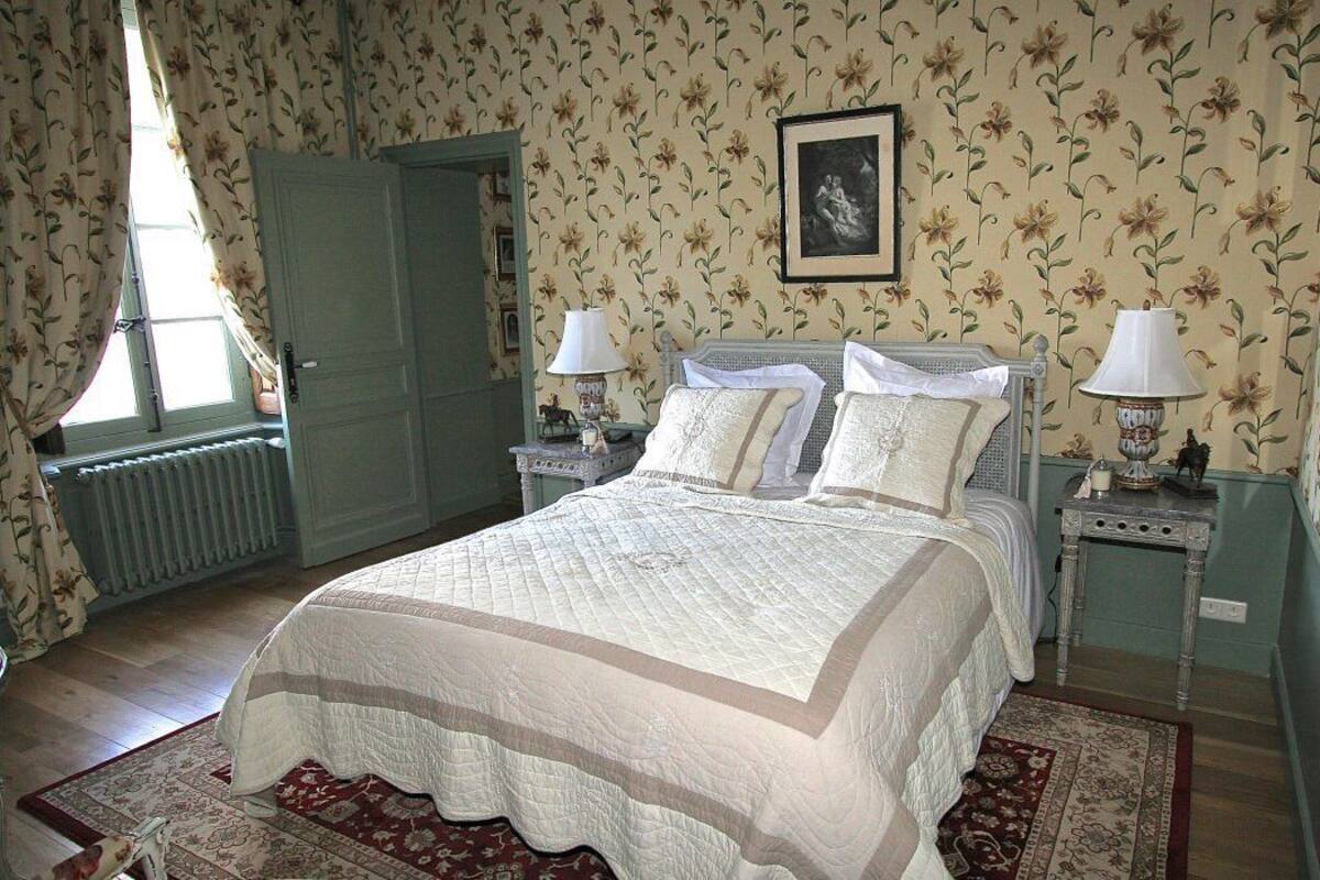 A bedroom with a large bed, two bedside tables with lamps, floral wallpaper, a radiator, and a door.