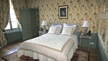 Un dormitorio con una cama grande, dos mesitas de noche con lámparas, papel tapiz floral, un radiador y una puerta.