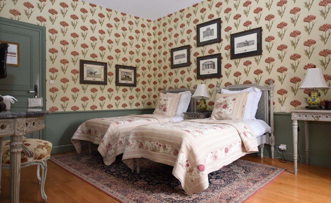 A bedroom with floral wallpaper, a double bed, two bedside tables with lamps, and a small table with a chair.