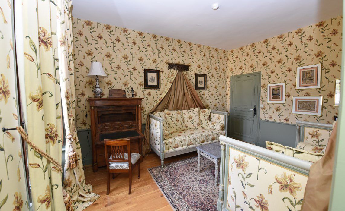 Quarto com papel de parede floral, cama com dossel, cómoda de madeira, cadeira e quadros na parede.