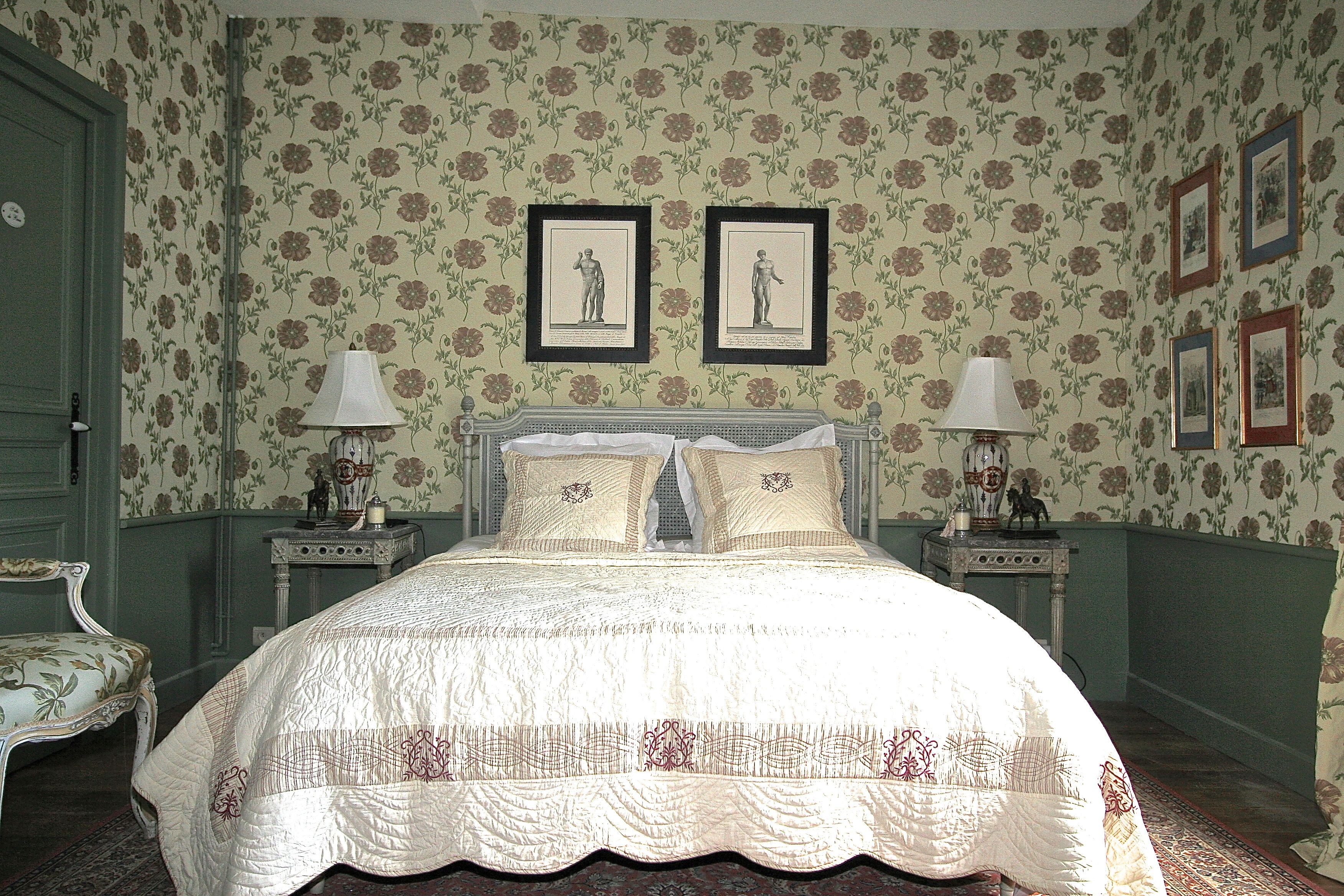 Ein klassisches Schlafzimmer mit floralem Wandpapier, einem Himmelbett, zwei gerahmten Bildern über dem Bett und einem Nachttisch mit einer Lampe auf jeder Seite.