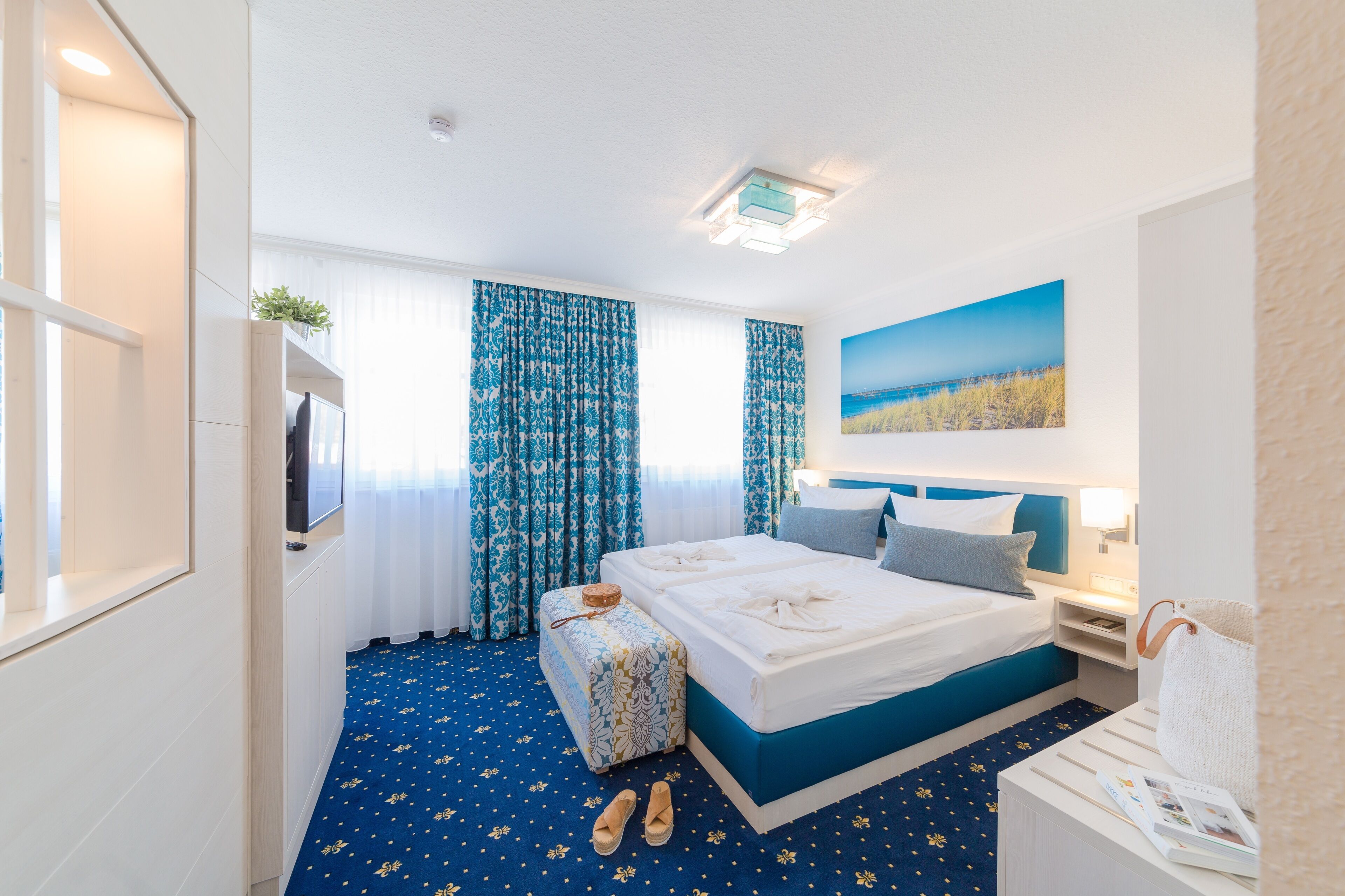 Una habitación de hotel con una cama grande, una mesita de noche con lámpara, una ventana con cortinas y un cuadro en la pared.