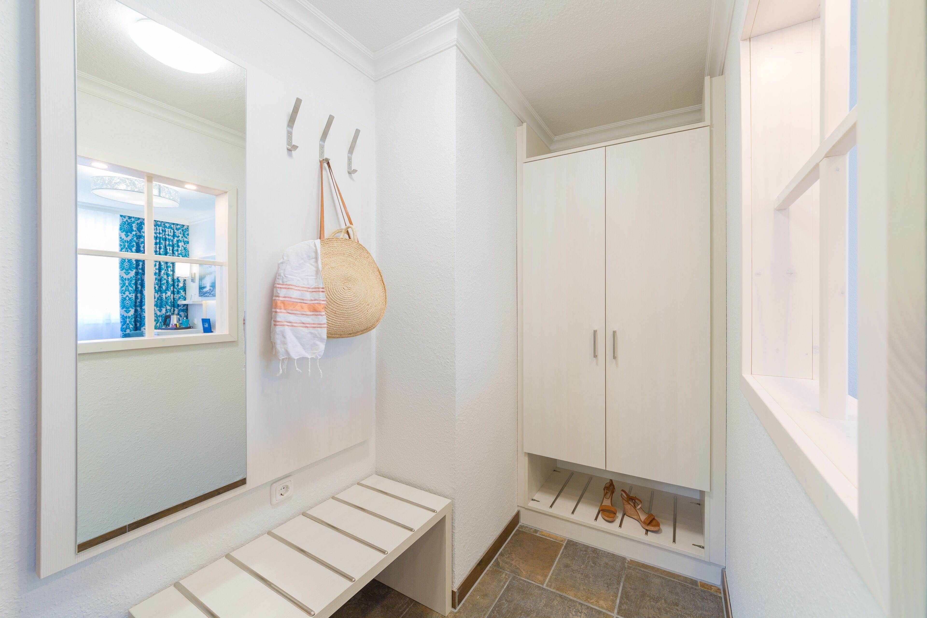 Une salle de bain avec un grand miroir, un porte-serviettes, une étagère où sont rangées des chaussures et une fenêtre donnant sur une pièce aux motifs bleus.