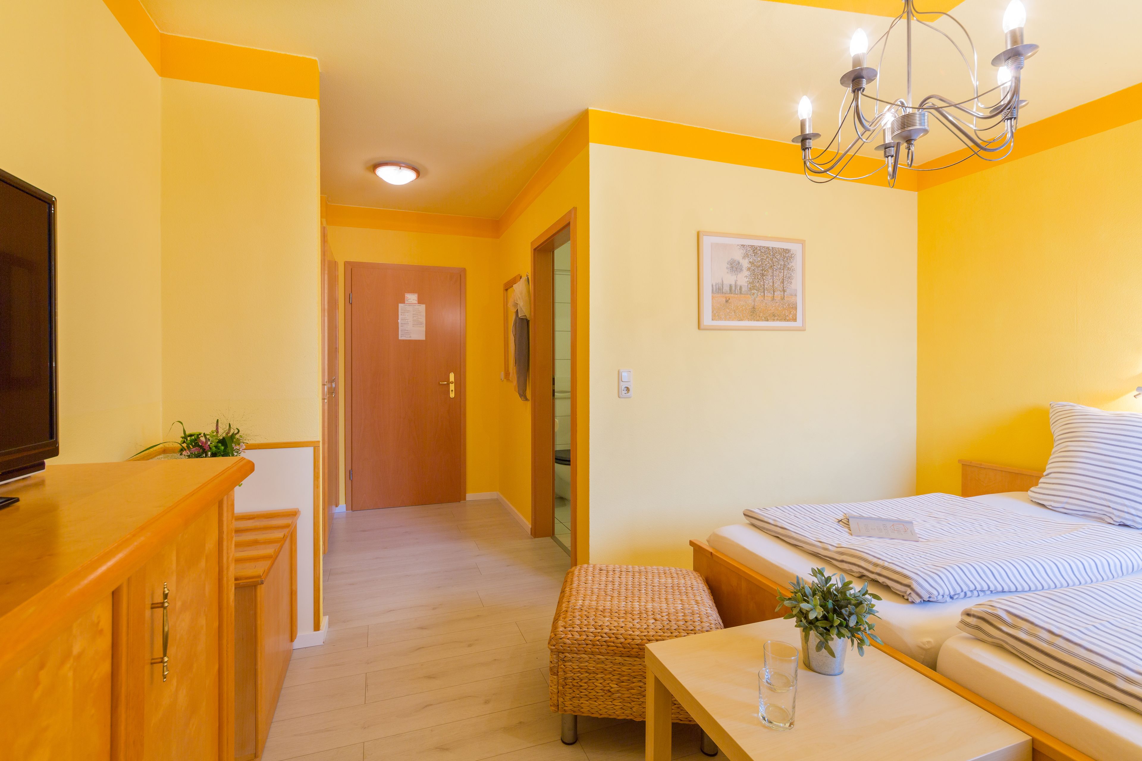 Una camera d'albergo con una scrivania in legno, un letto con lenzuola a righe, un piccolo tavolino con una pianta e un lampadario.