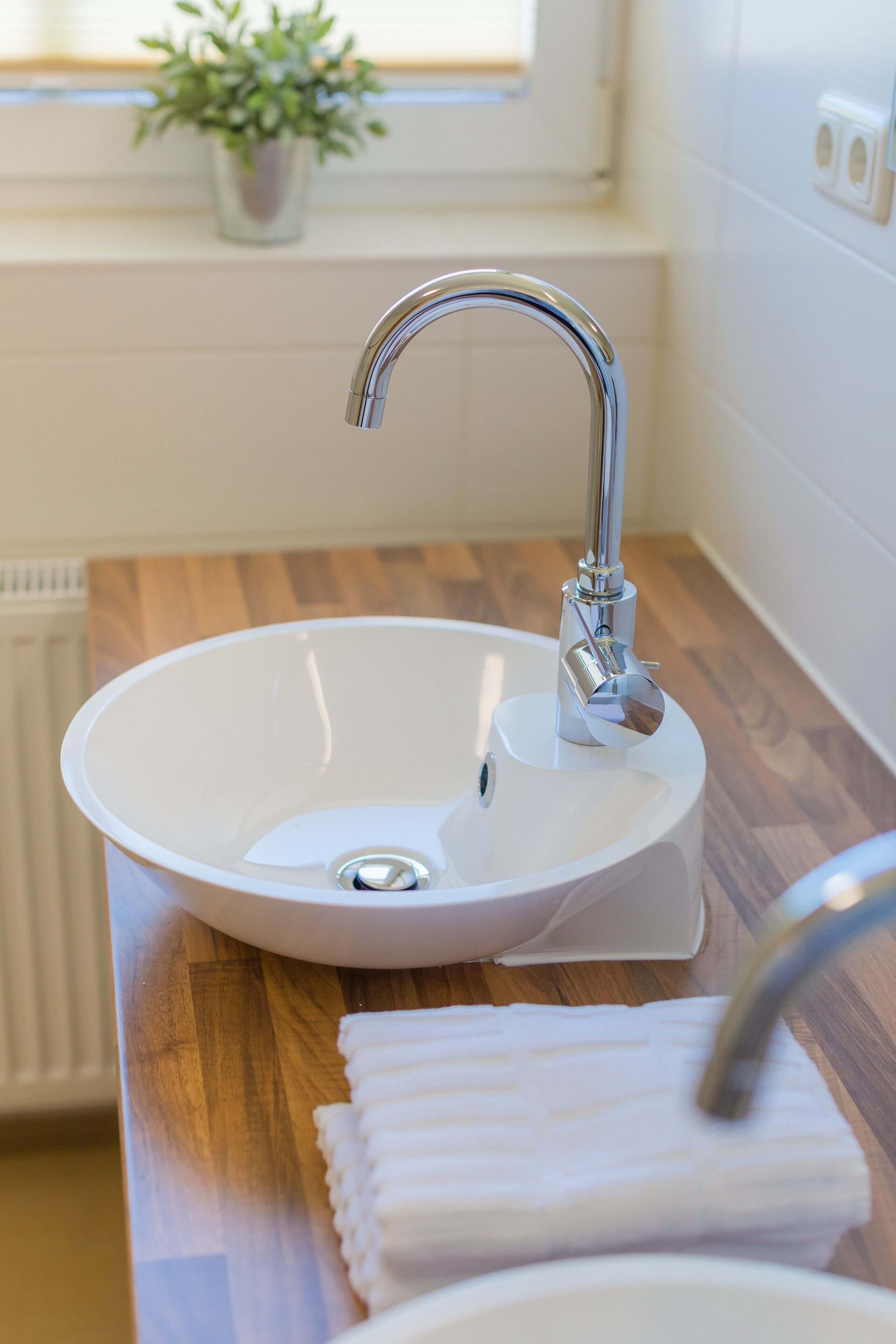 A white sink with a chrome faucet in a bathroom with wooden flooring and a window with a plant.