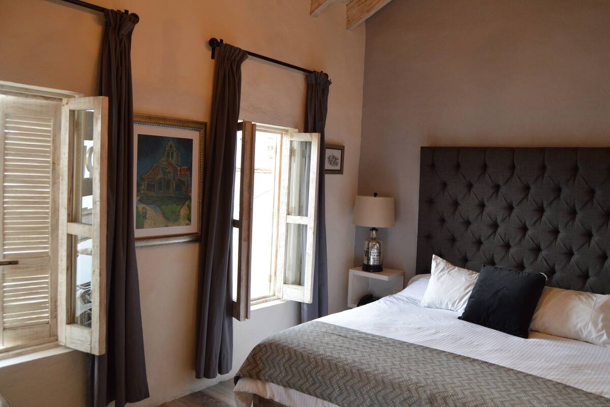 A bedroom with a large bed, a nightstand with a lamp, a window with curtains, and a painting on the wall.