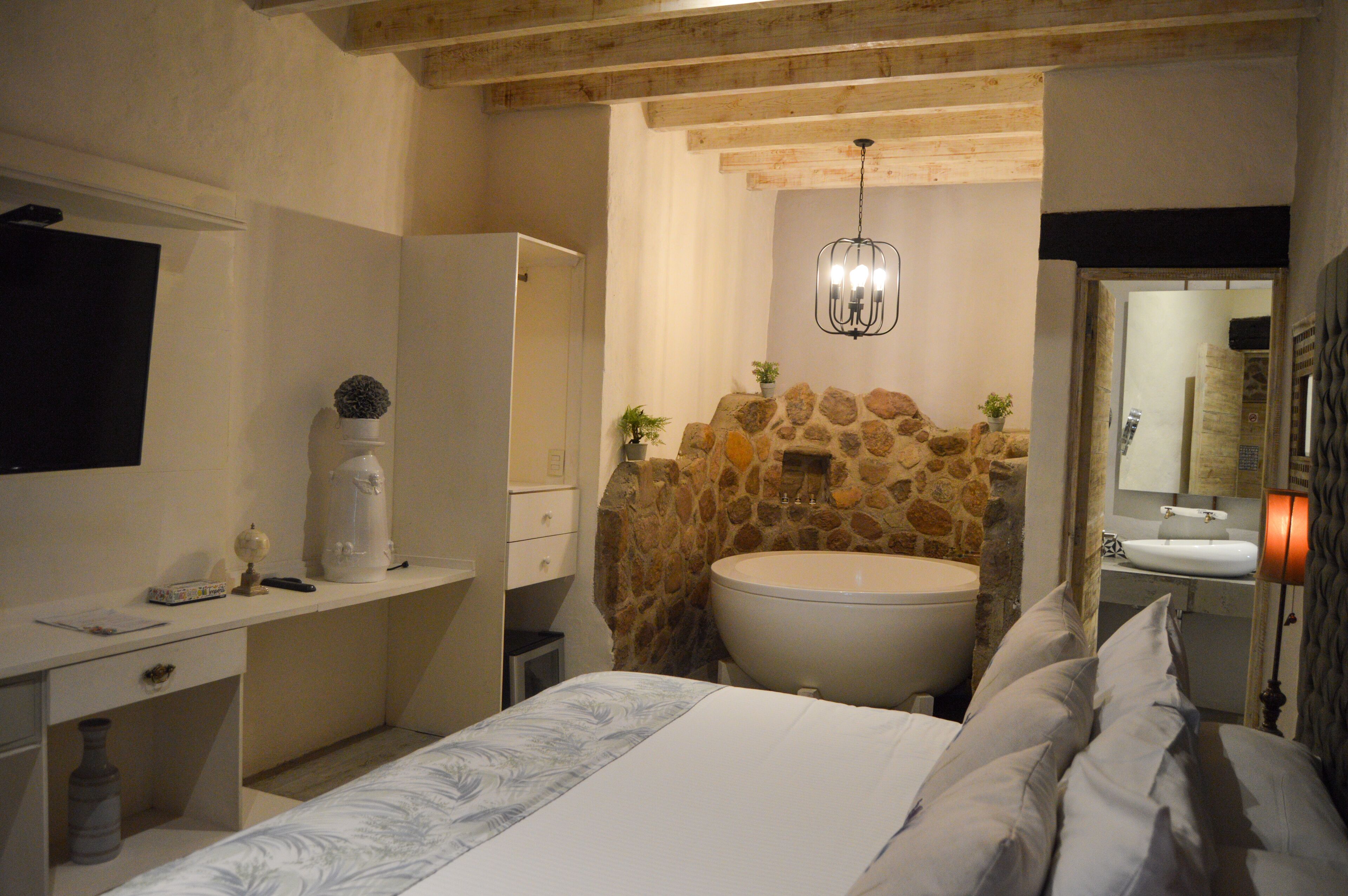 A bedroom with a large bed, a bedside table, a dresser, a vanity with a mirror, a stone accent wall, and a bathtub.