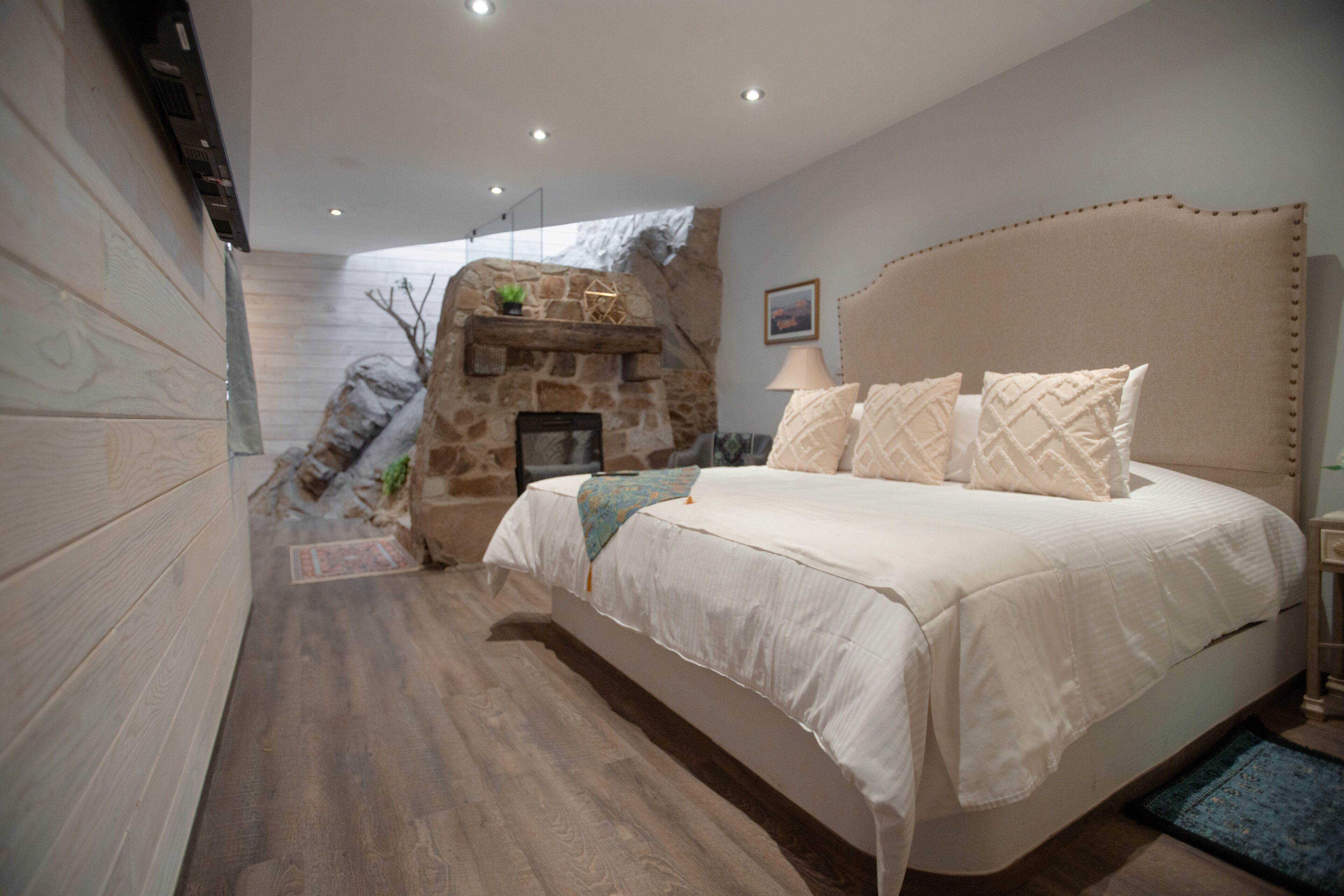Une chambre avec une cheminée en pierre, un grand lit doté d’une tête de lit et un plancher en bois.