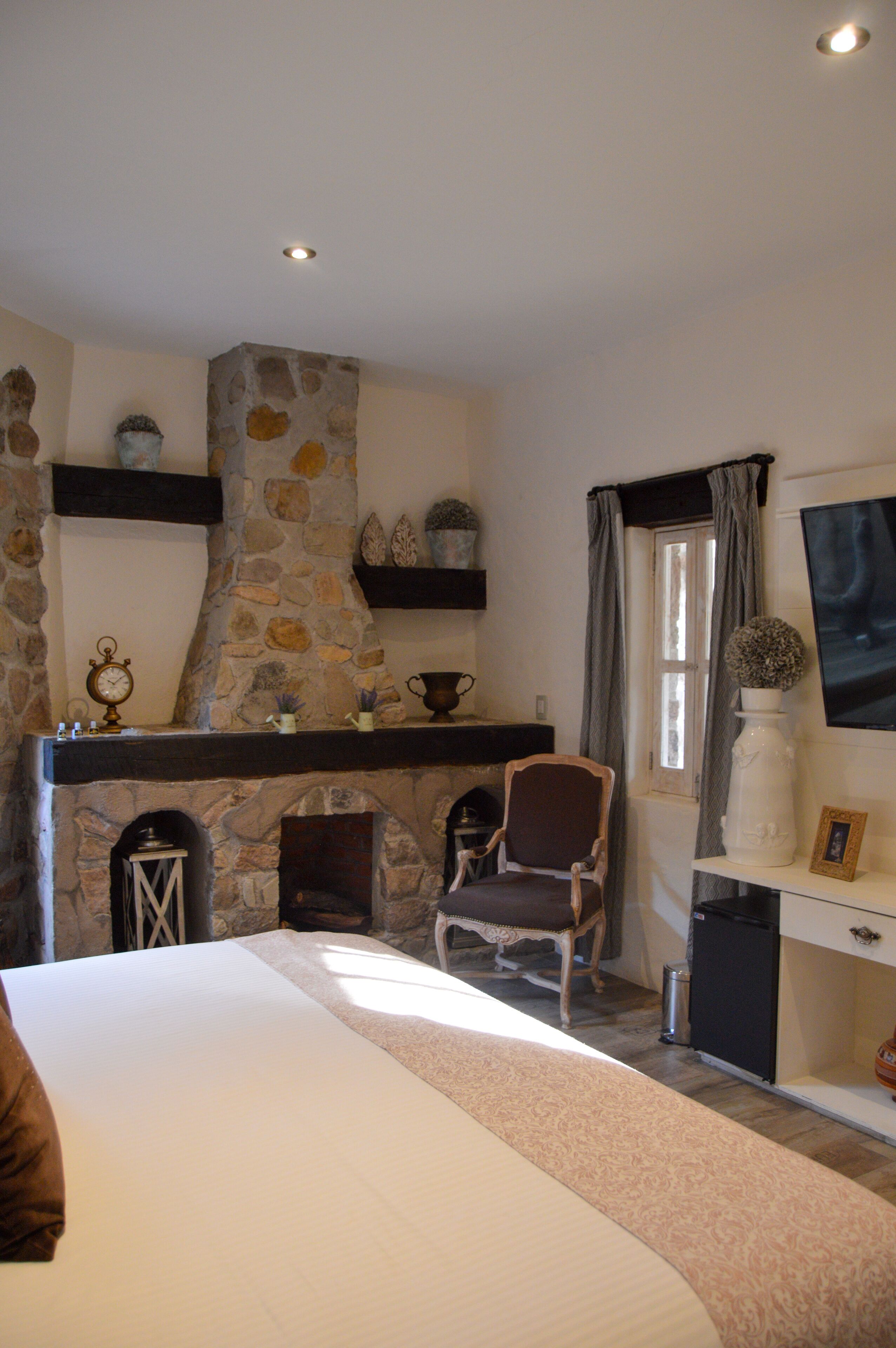 A bedroom with a stone fireplace, a bed, a chair, and a desk with a vase and picture frame.