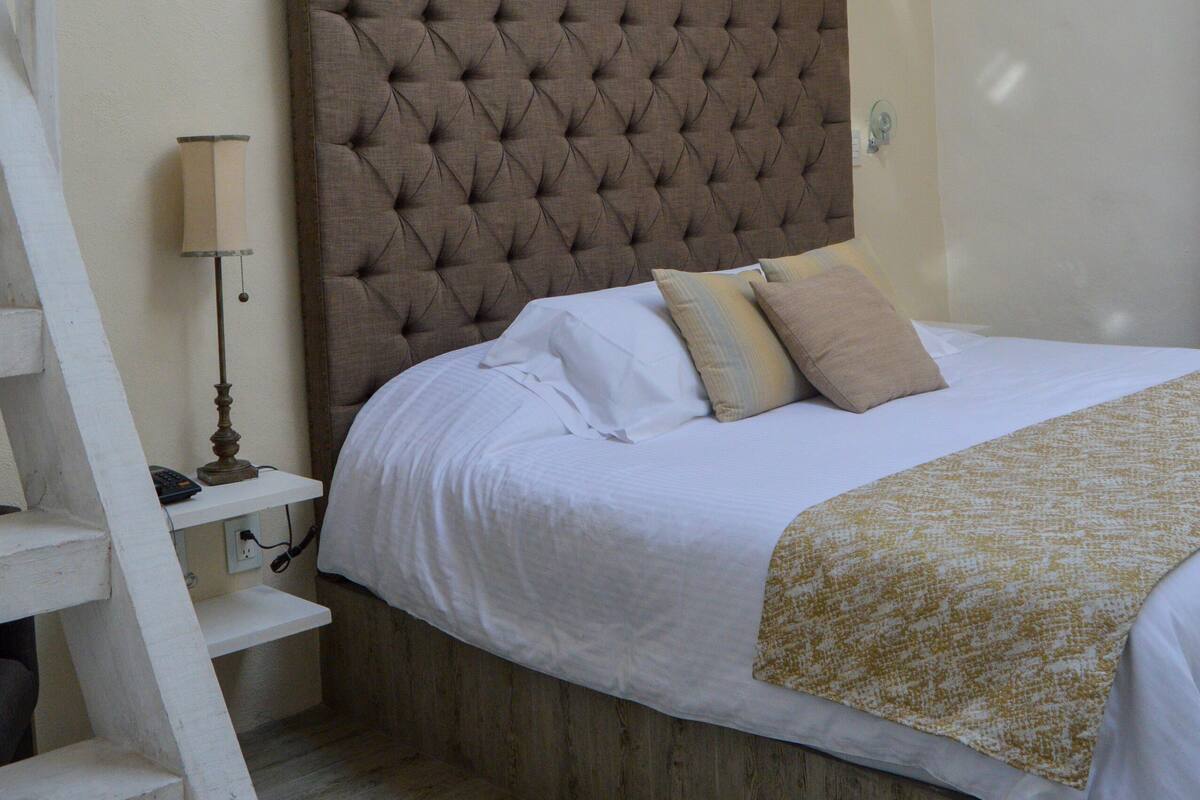A bedroom with a large tufted headboard, a wooden bed frame, a white bedspread, and a wooden floor.