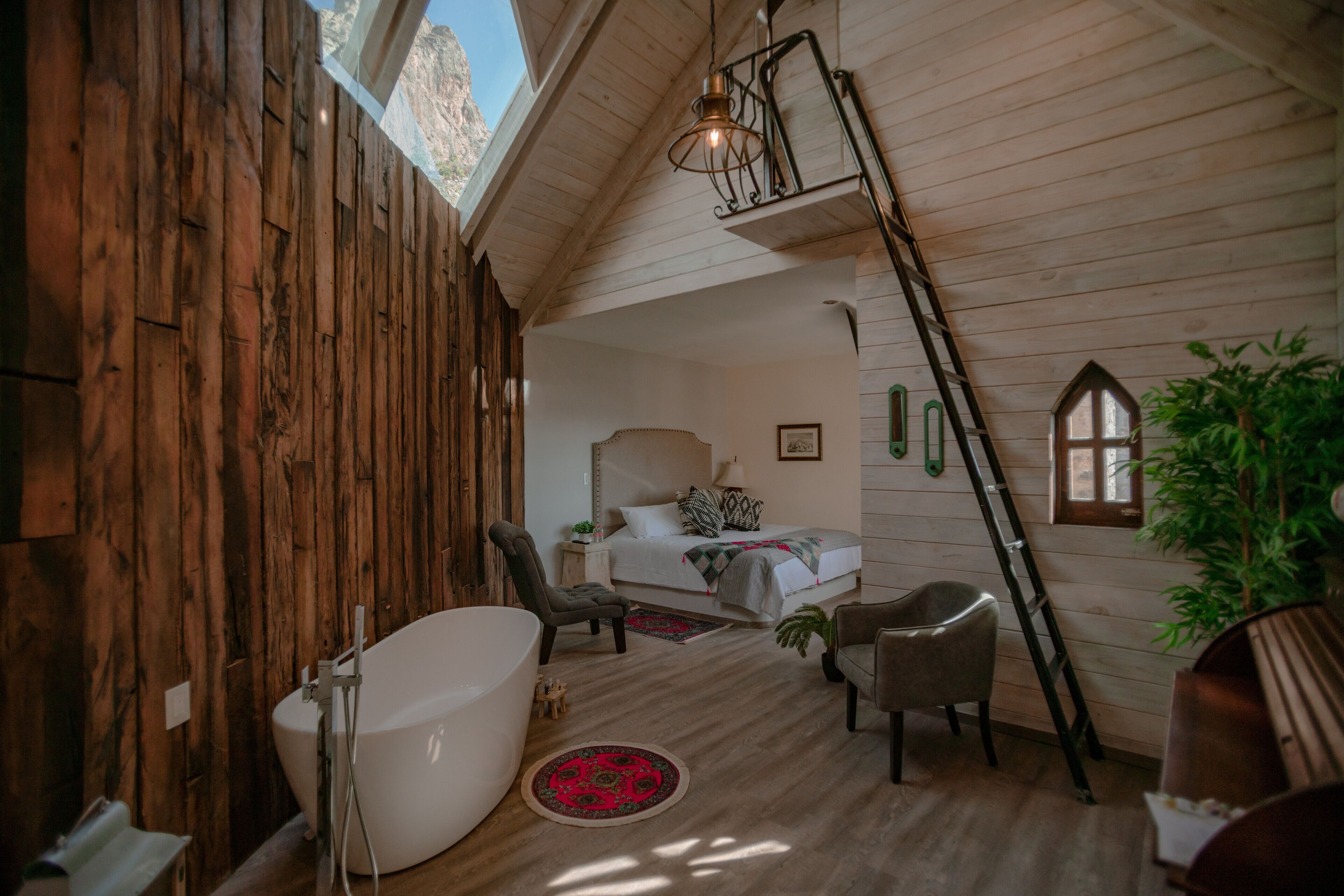 Een rustieke slaapkamer met een houten plafond, een groot bed, een zithoek met een stoel, een badkuip en een ladder naar het slaapgedeelte boven.