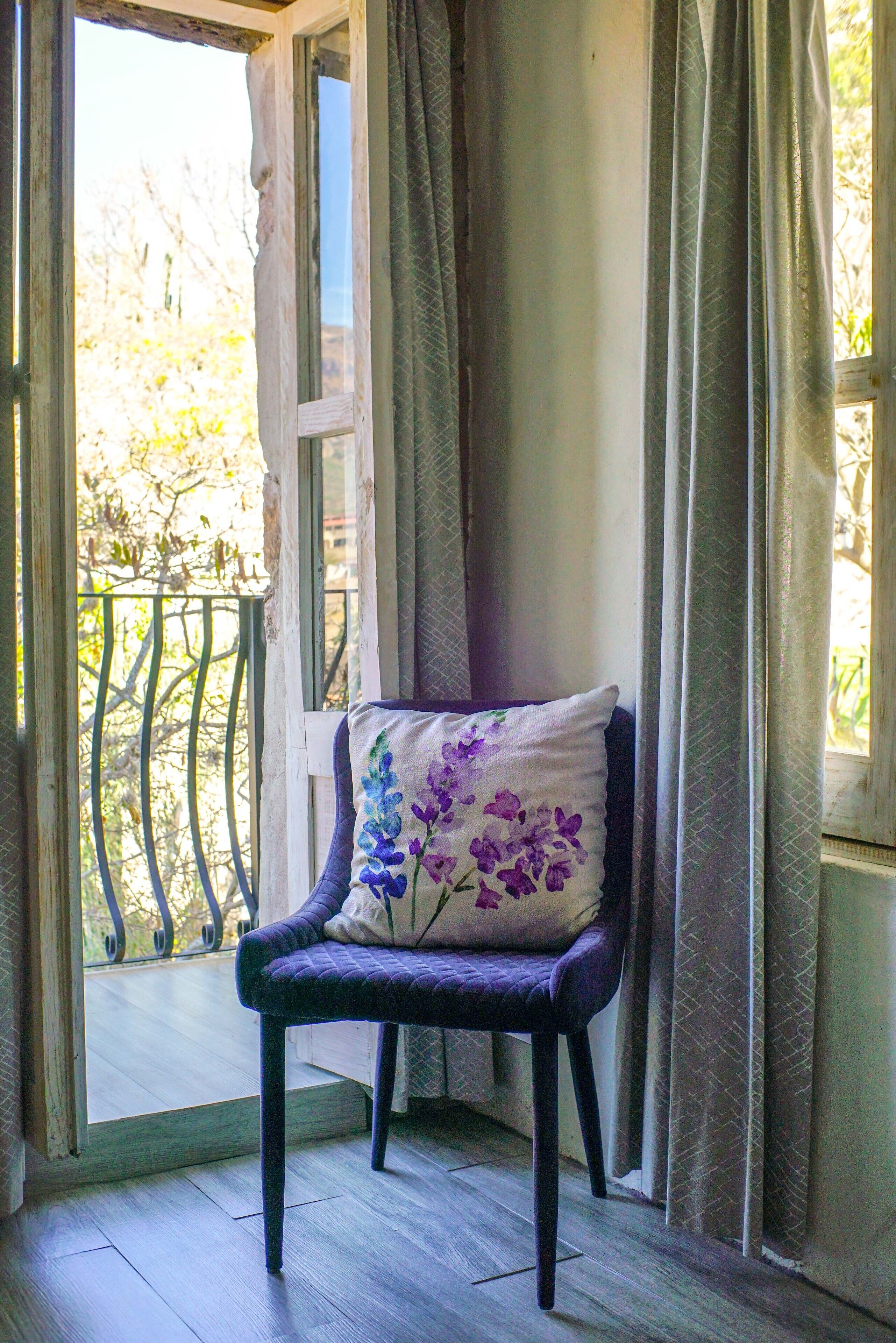 A chair with a floral pillow in a room with a view through a window with curtains.