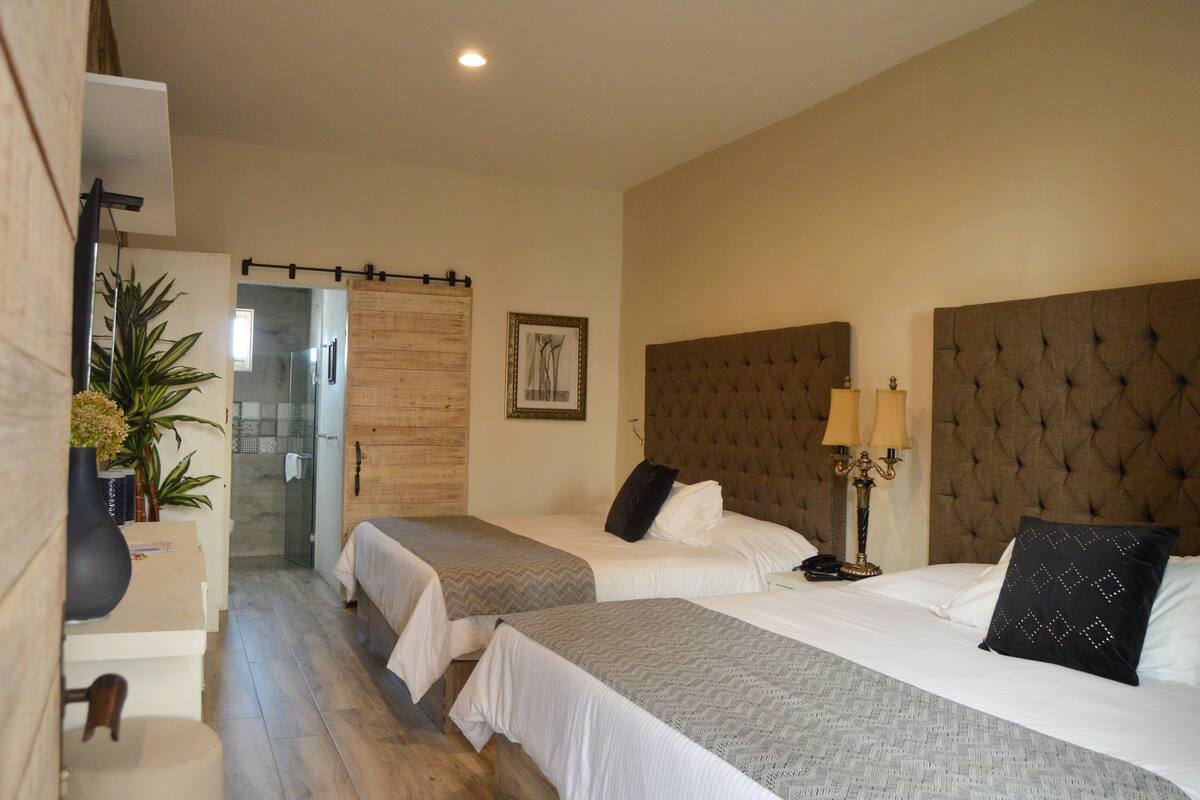 A hotel room with two beds, a wooden door, a framed picture, and a bedside lamp.