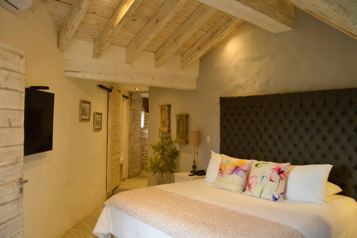 A bedroom with a large bed, wooden ceiling beams, a stone wall, and a television mounted on the wall.
