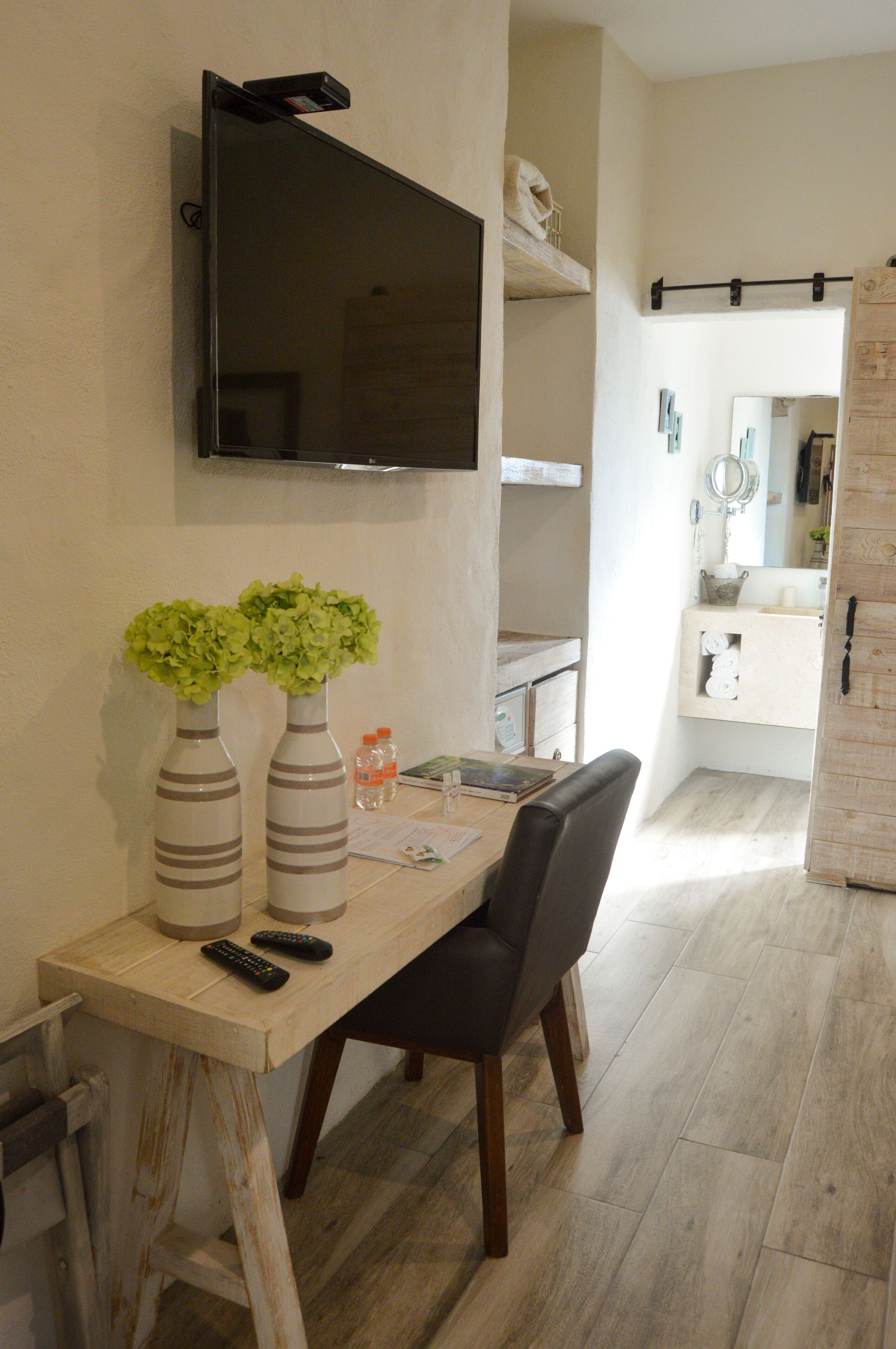 Un comedor con una mesa, sillas, un televisor montado en la pared y dos jarrones con flores verdes.