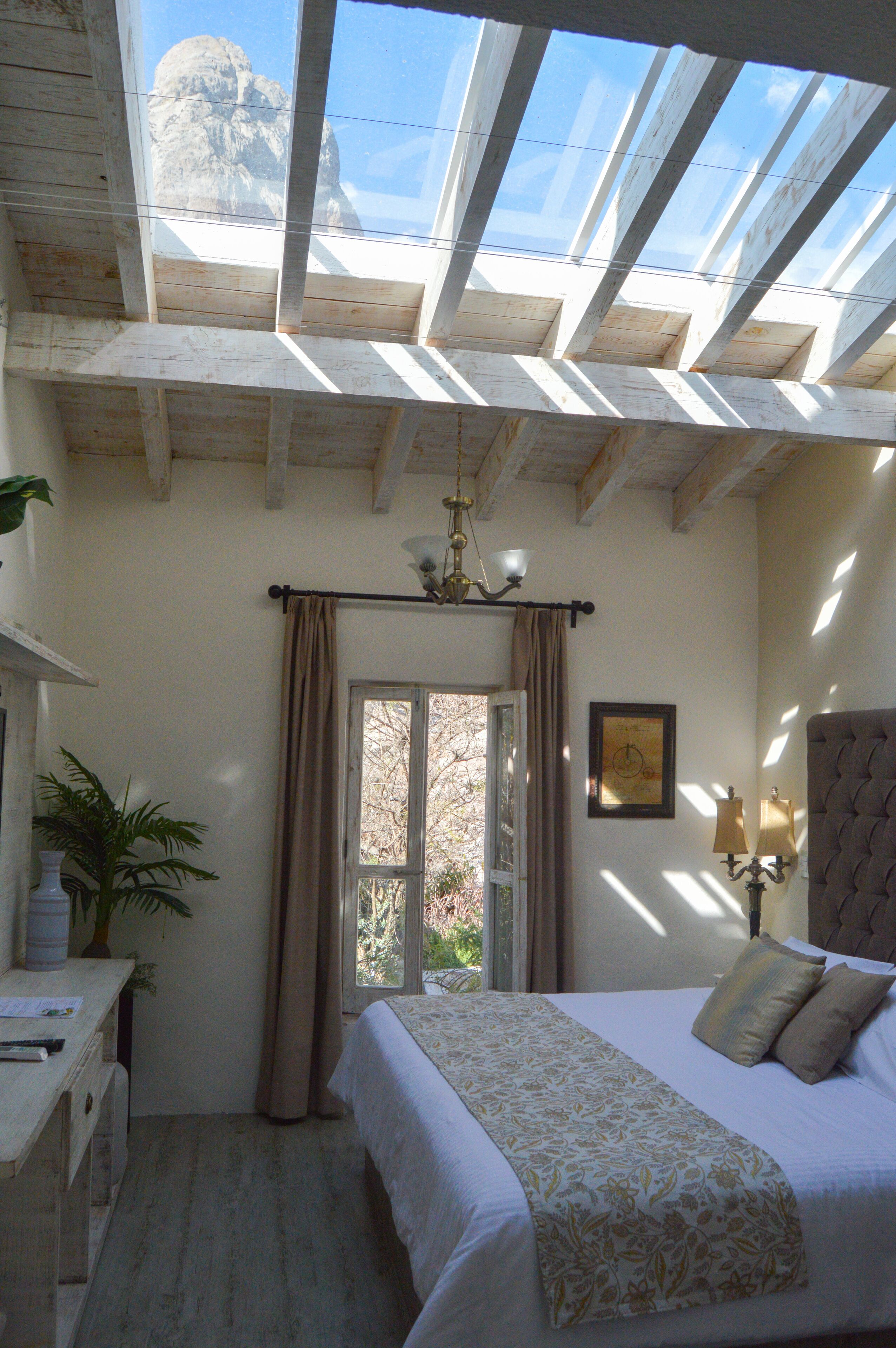 A bedroom with a skylight, a bed with a floral bedspread, a bedside table, a lamp, and a window with curtains.