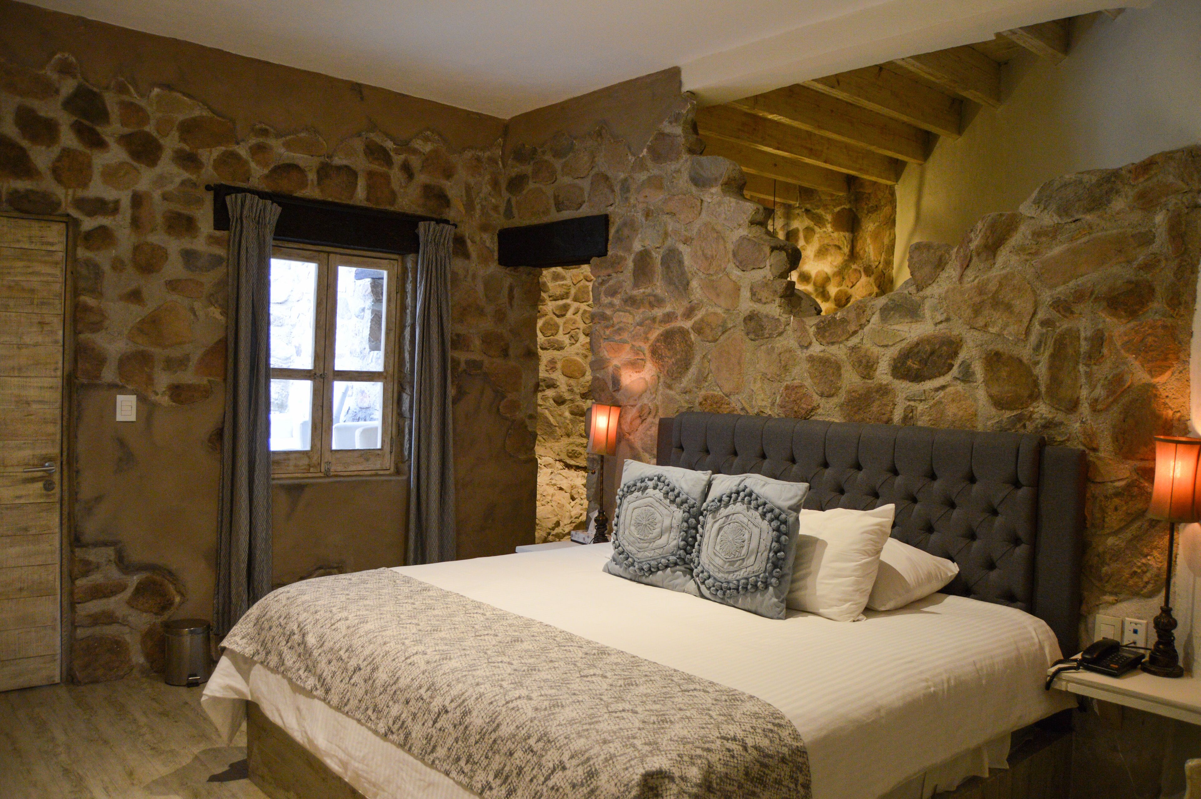 A bedroom with a stone wall, a bed with a grey upholstered headboard, and a window with curtains.