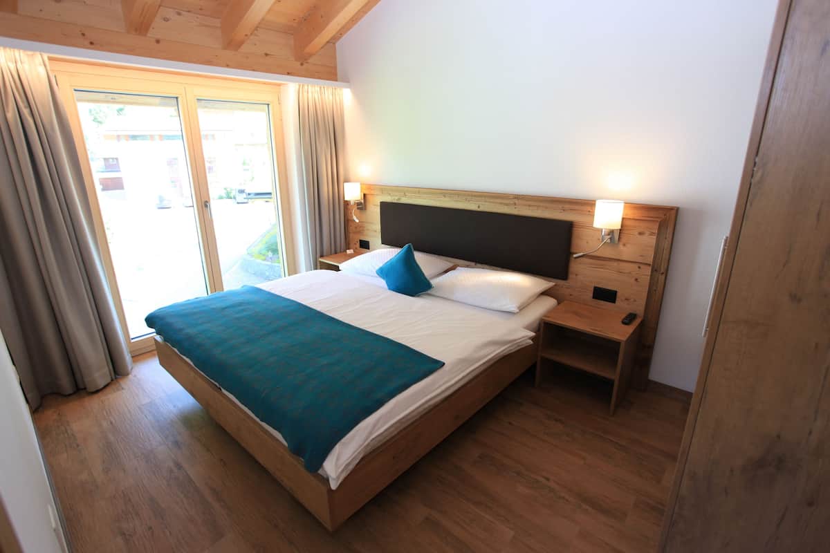 Una cama con estructura de madera, cubierta con un edredón verde azulado, sábanas blancas y una almohada azul. Hay dos mesitas de noche con lámparas. El cuarto tiene piso de madera y un ventanal con cortinas.