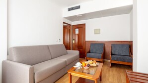 Un salón con un sofá color beige, dos sillones, una mesa de centro de madera, y una mesita con frutas y una tetera.