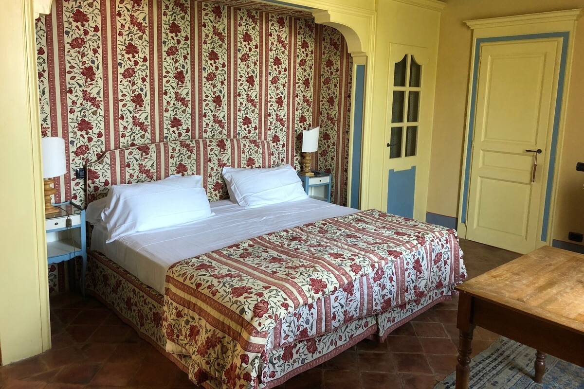 A bedroom with a floral-patterned bedspread, a wooden nightstand with a lamp, and a door with a blue trim.