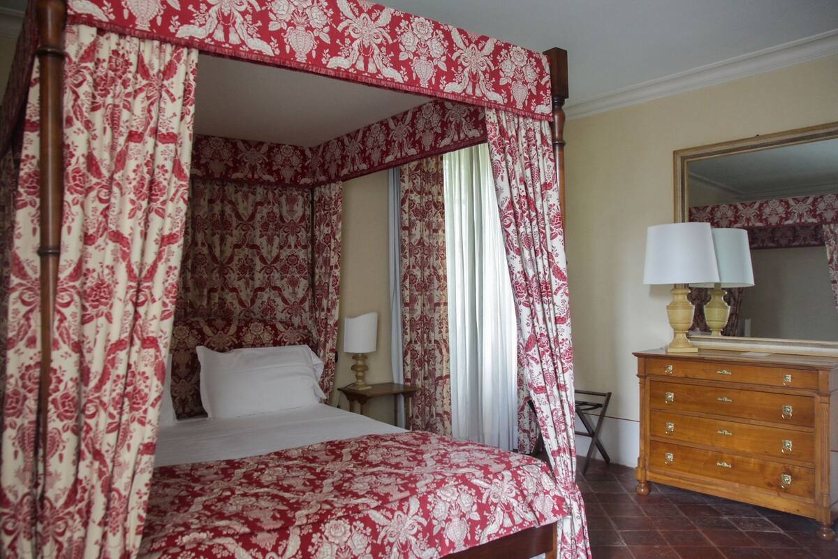 A four-poster bed with a floral canopy, a wooden dresser with drawers, a mirror, and a nightstand with a lamp.