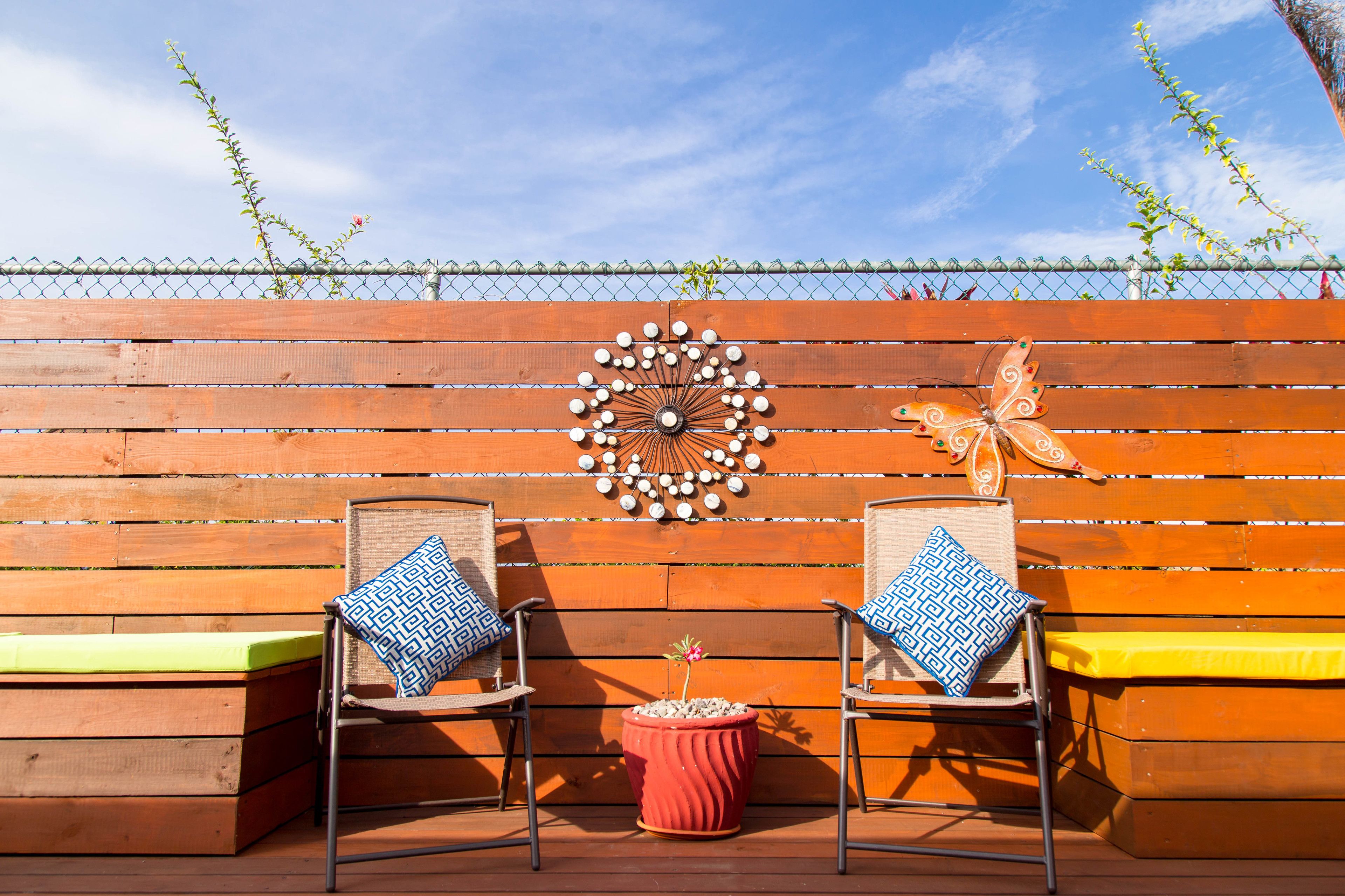 A wooden deck with two chairs and a bench, a decorative wall with a sunburst and butterfly, and a red pot with a plant.