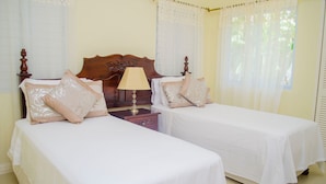 Quarto de hotel com duas camas de solteiro, cabeceira de madeira, mesa de cabeceira com abajur e cortinas de renda.