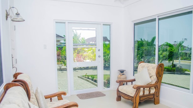 Quarto com cadeira de madeira, sofá, porta de vidro deslizante grande que dá para um jardim e uma pequena mesa com um vaso.