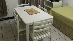A small, clean room with a white table and chairs, a television on a stand, and a bed with a green cover.