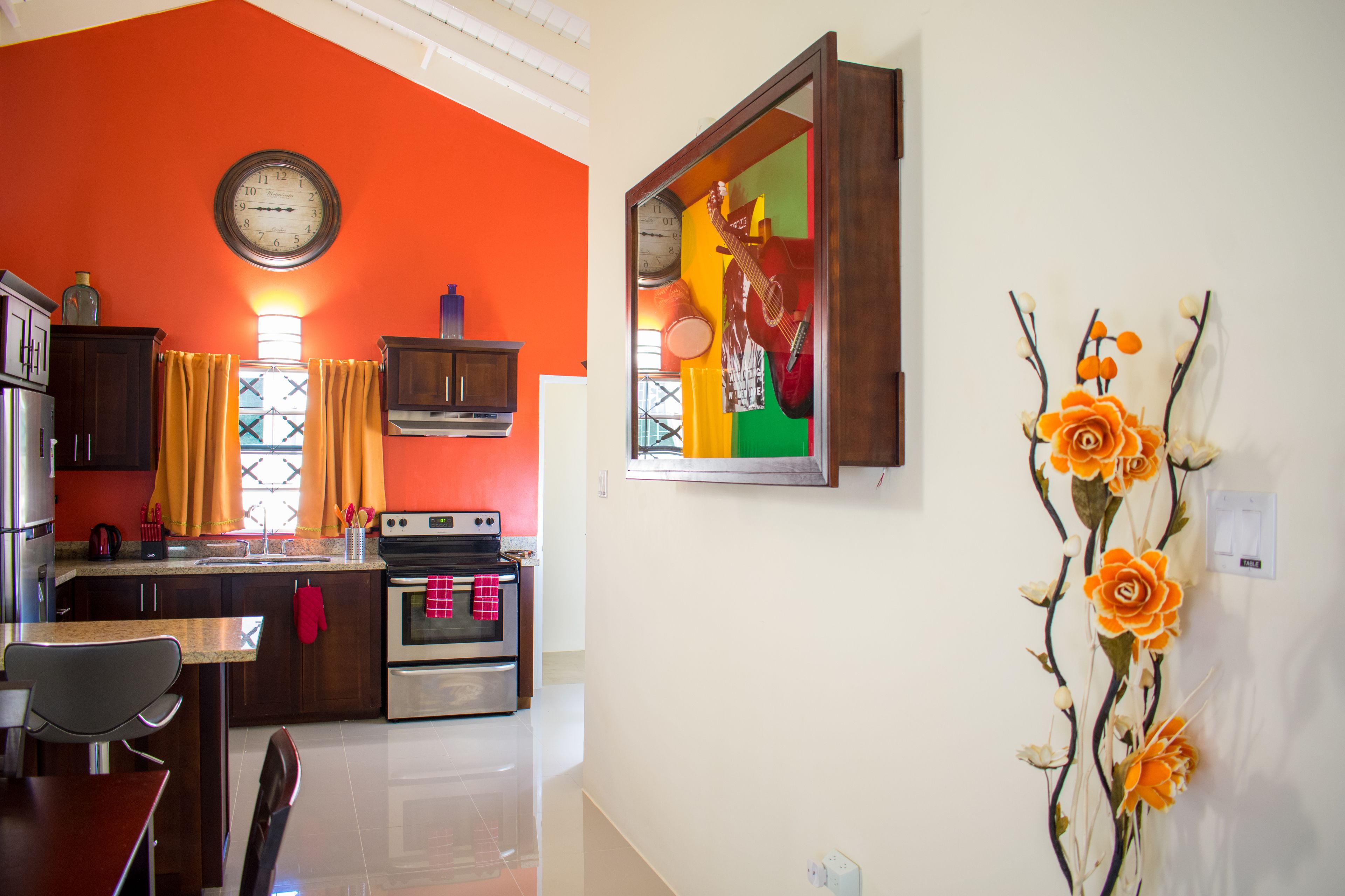 A kitchen with a clock, a stove, and a dining area with chairs.