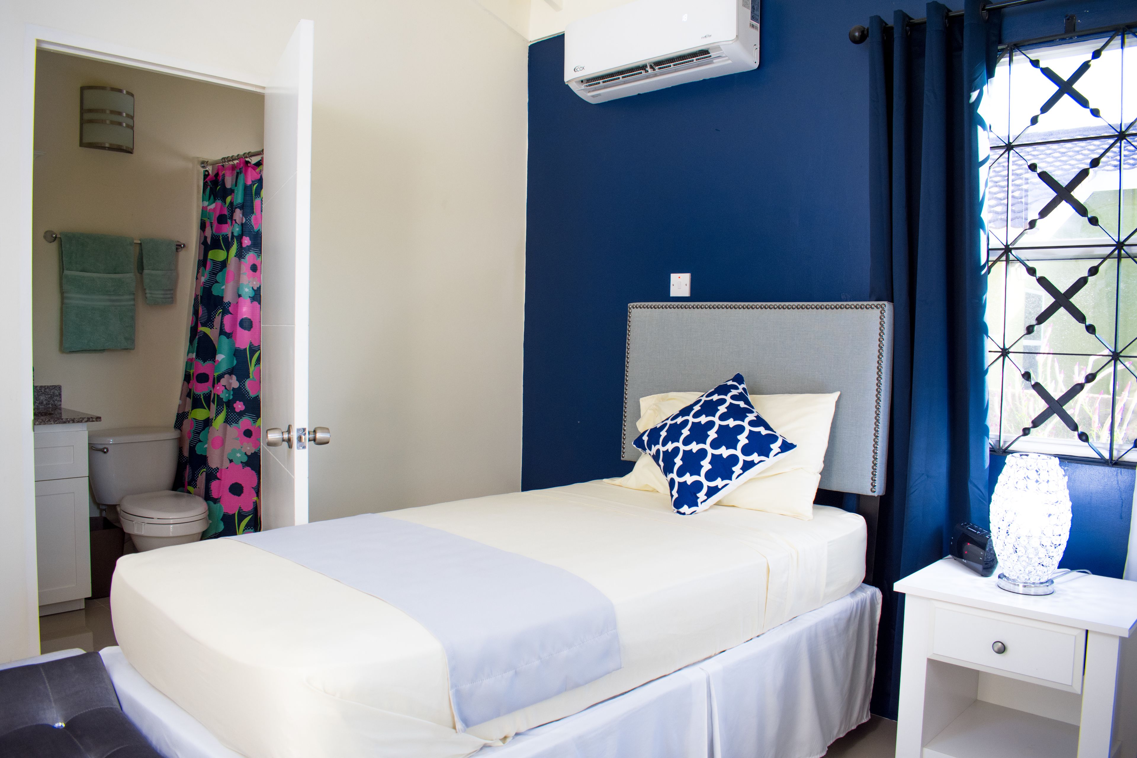 A bedroom with a bed, a bedside table with a lamp, a window with curtains, and an air conditioning unit.