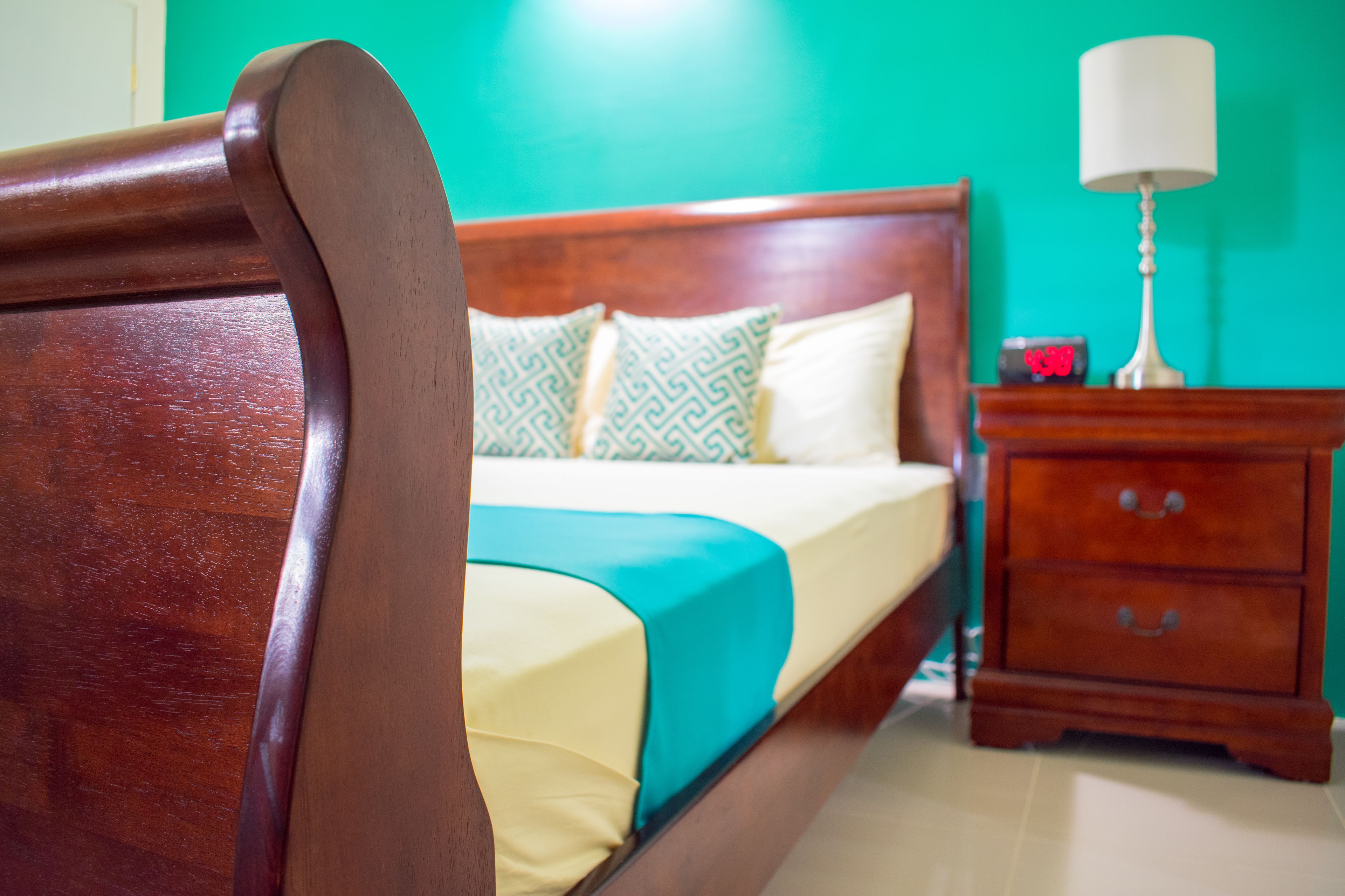 Een houten bed met een deken in teal- en geeltinten, een houten nachtkastje met een lamp, en een groene muur.