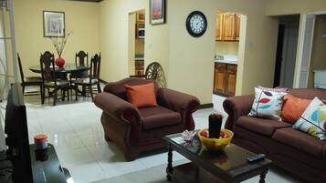 A living room with two brown sofas, a coffee table with a bowl of fruit, a TV, and a dining area with a table and chairs.