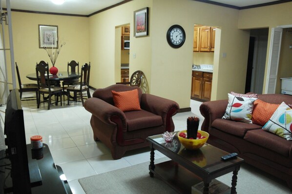 A living room with two brown sofas, a coffee table with a bowl of fruit, a TV, and a dining area with a table and chairs.