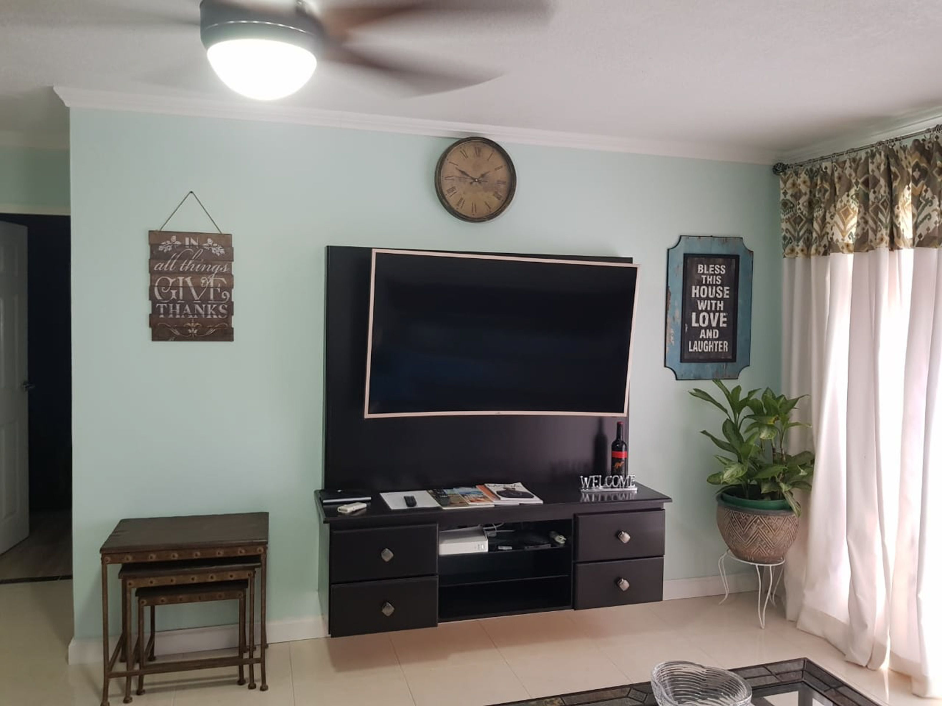 A living room with a flat-screen TV, a clock, a 'WELCOME' sign, a small table, and a potted plant.