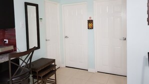 Quarto com duas portas brancas, uma pequena mesa com cadeiras, um espelho e um aparelho de iluminação numa das portas.